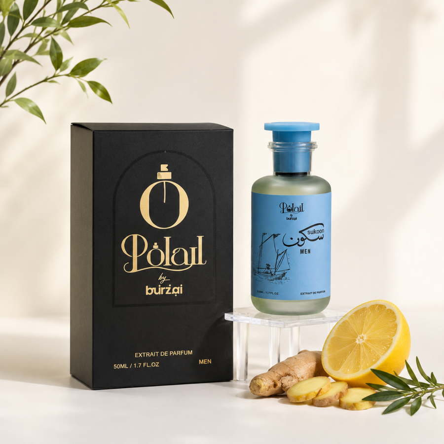 A bottle of sukoon perfume for men with a light blue label and blue cap, displayed next to its black packaging box with gold lettering. The setup is styled with fresh ginger roots, a sliced yellow lemon, and green leaves on a white surface with soft shadows.